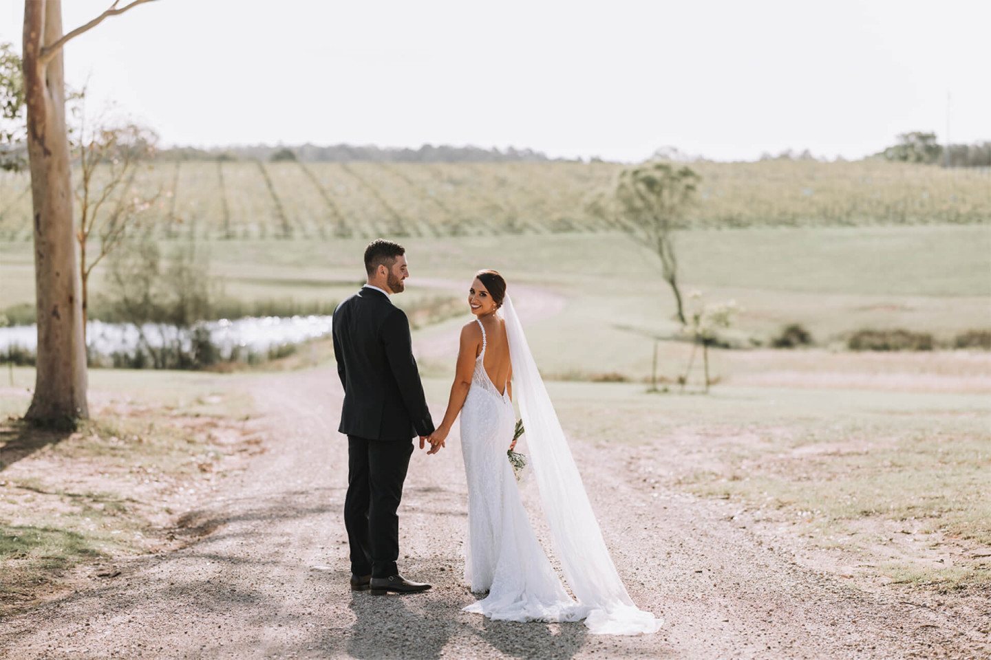 A couple on the white gravel drive at Squire&rsquo;s, the vineyard and dam behind